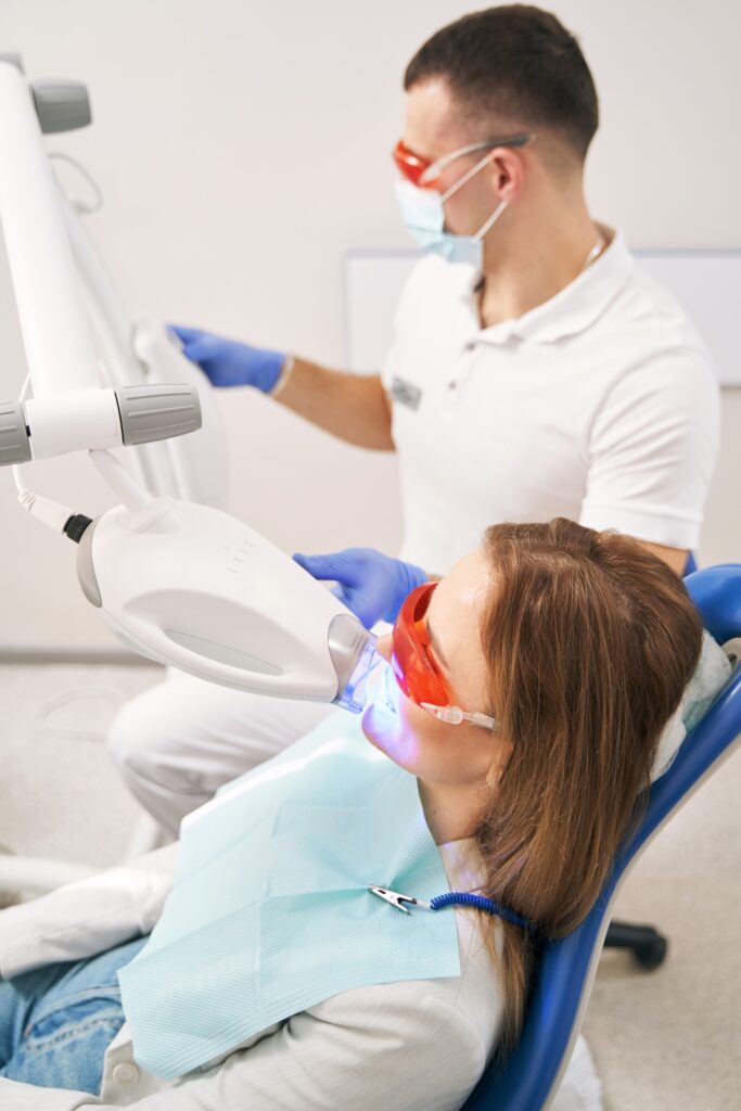 Female patient in protective glasses sitting in dental chair while dentist performing dental treatment with laser device